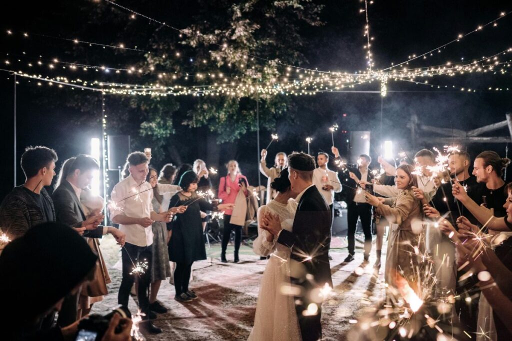 A couple celebrate their wedding party together with guests. The guests celebrate with sparkler fireworks.