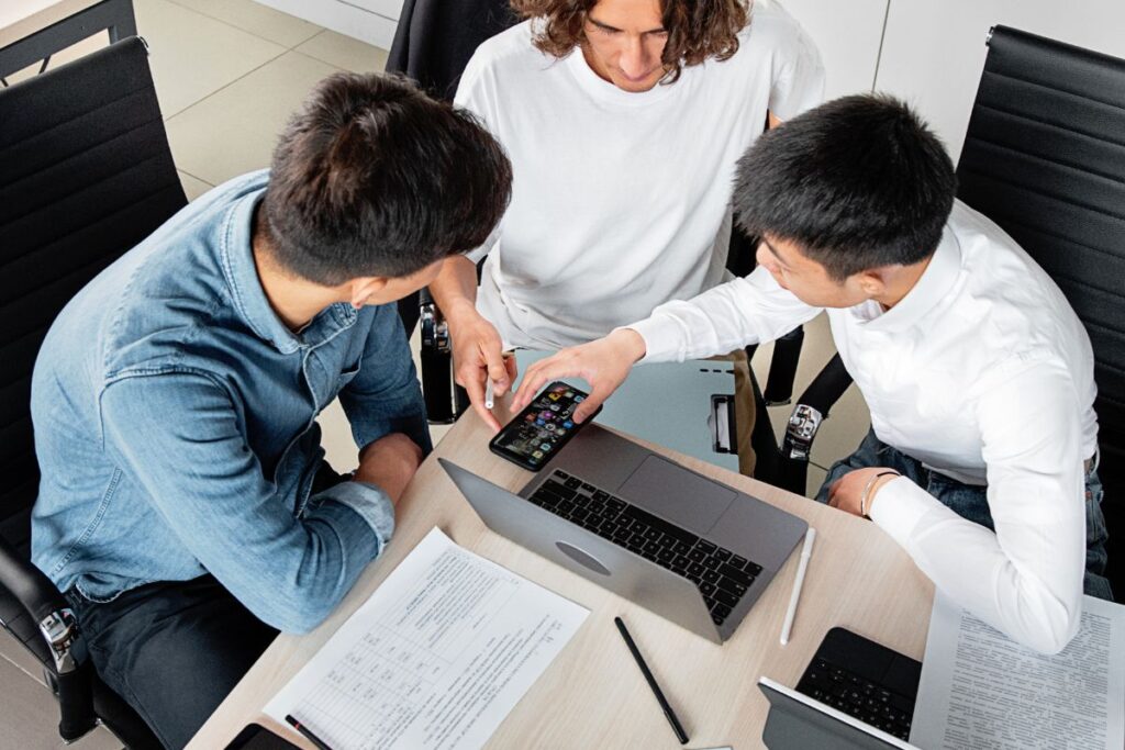 A group of three male employees sit at the end of a table and discuss work over a phone and laptop. There is a pile of paperwork on the table beside them