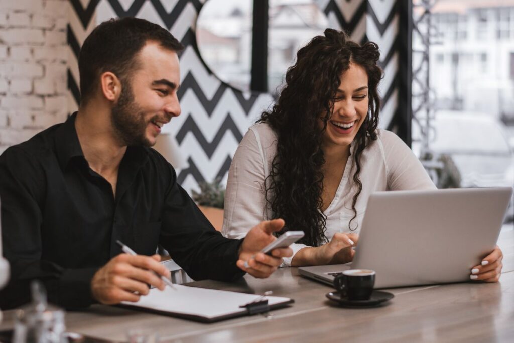 Two co-workers collaborate together during a meeting in a café. There is coffee on the table alongside a laptop and paperwork. They are smiling and laughing together. 