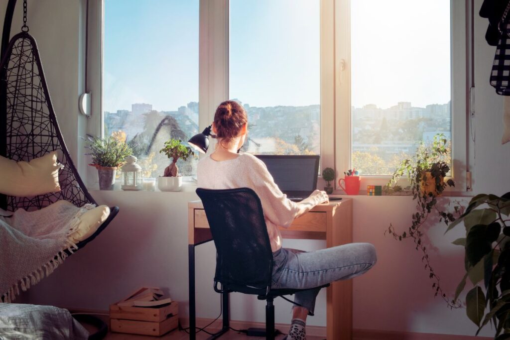 A woman works remotely at her desk in her bedroom. She sits by the window. It is a sunny day. There are house plants on the window ledge in front of her.