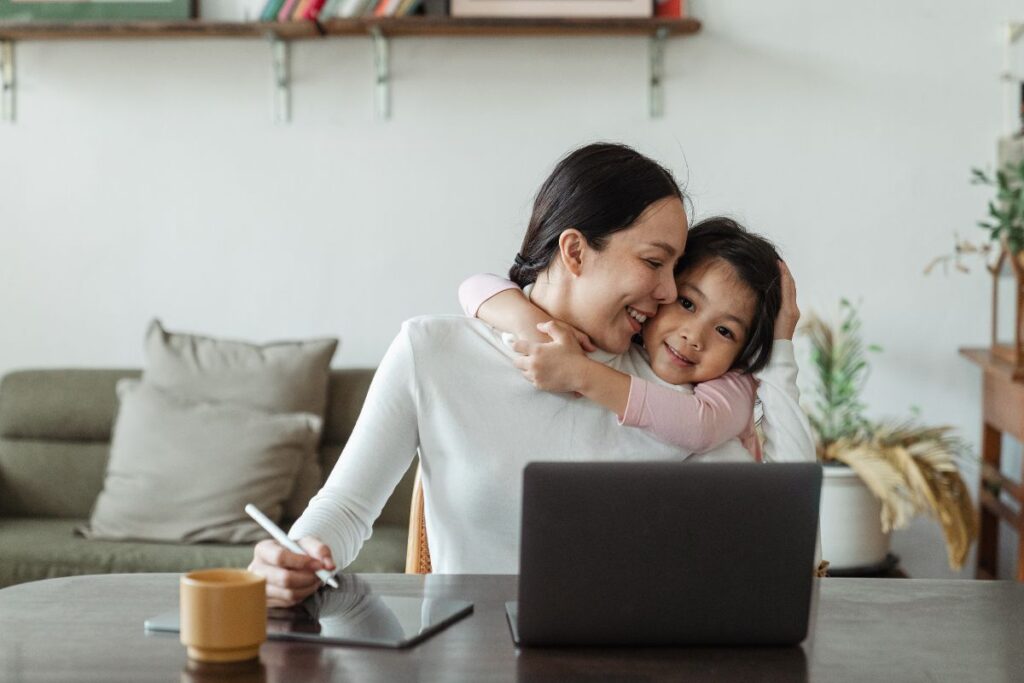 A woman works from home and hugs her daughter at the same time. 