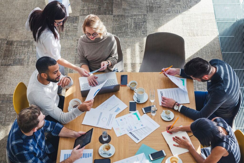 A birds-eye perspective photo of a team work together in a meeting. There is lots of paperwork on the table, and the team take part in a discussion. Some take notes and some scroll on tablets.