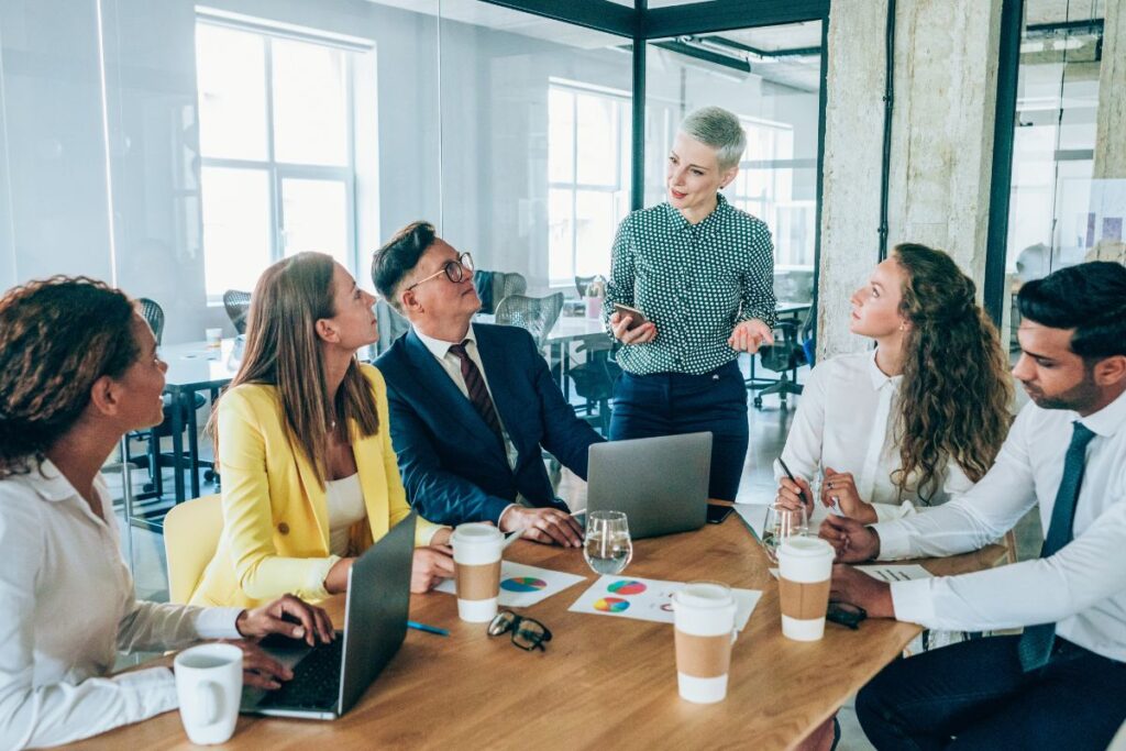 A manager runs a meeting with her team. She stands up at the end of a table where five team members sit, drink coffee, make notes, and listen.