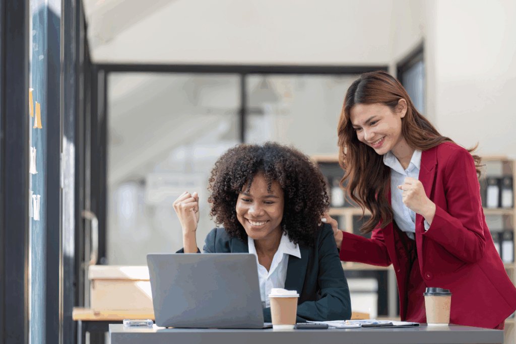 Two female co-workers celebrate at their desks. There is a laptop on the table along with two coffees.