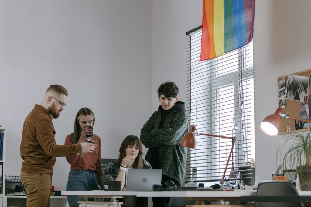 A group of four employees look over one laptop as they collaborate as a team. They drink coffee; There is a rainbow flag hanging on the window