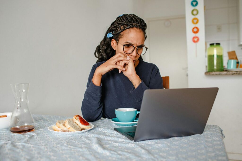 A woman works from home at her kitchen table. She has a cup of coffee in front of her, and her breakfast to the left of her. She has her laptop in front of her, and she has a concentrated expression on her face.