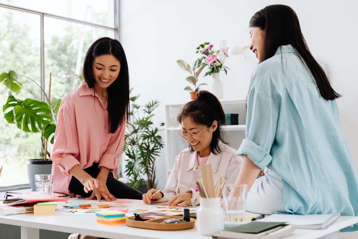 Three women work together on a project at work. Two sit on the desktop, and one sits on her desk chair in the middle. They are all smiling and having fun.