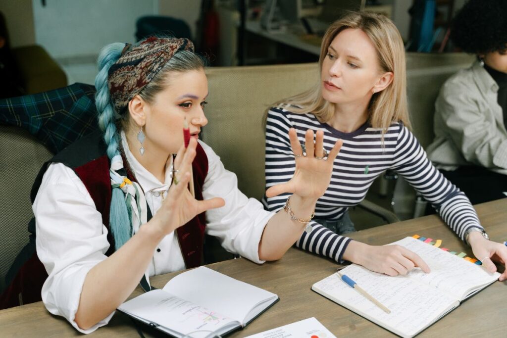 Two female employees sit together and creatively collaborate at work. One holds her hands up as she talks, while the other listens to what she has to say
