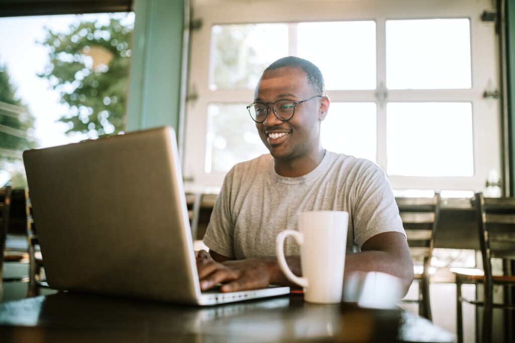 A man works remotely from a cafe. He smiles as he types on his laptop