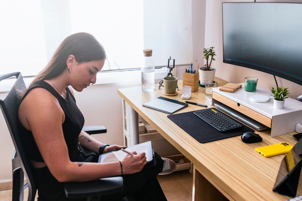 A woman works remotely at her desk. She holds a tablet on her lap and makes notes. There is a desktop computer in front of her, alongside a collection of houseplants.