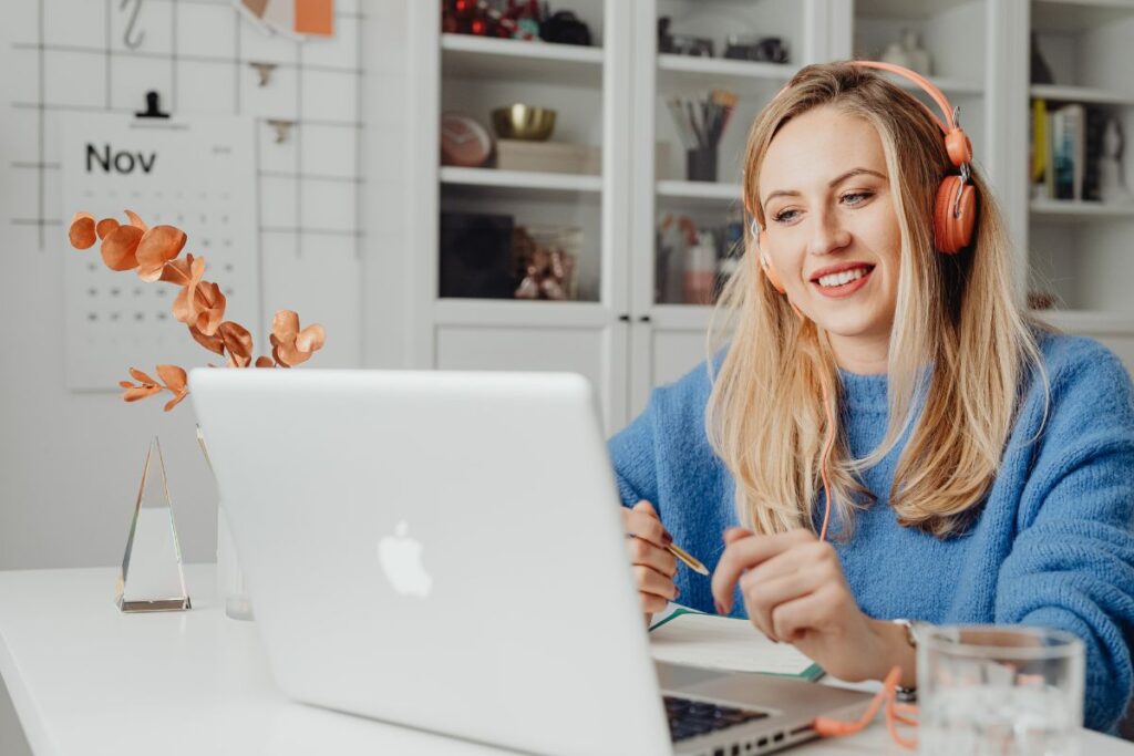 A woman sits on a remote meeting. She is wearing headphones in her kitchen. She smiles at her laptop.