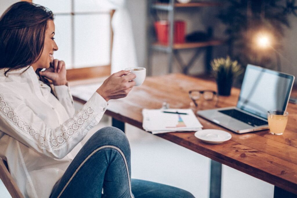 A woman works from home. She has her laptop and notes on her desk. She drinks coffee while she talks on the phone.