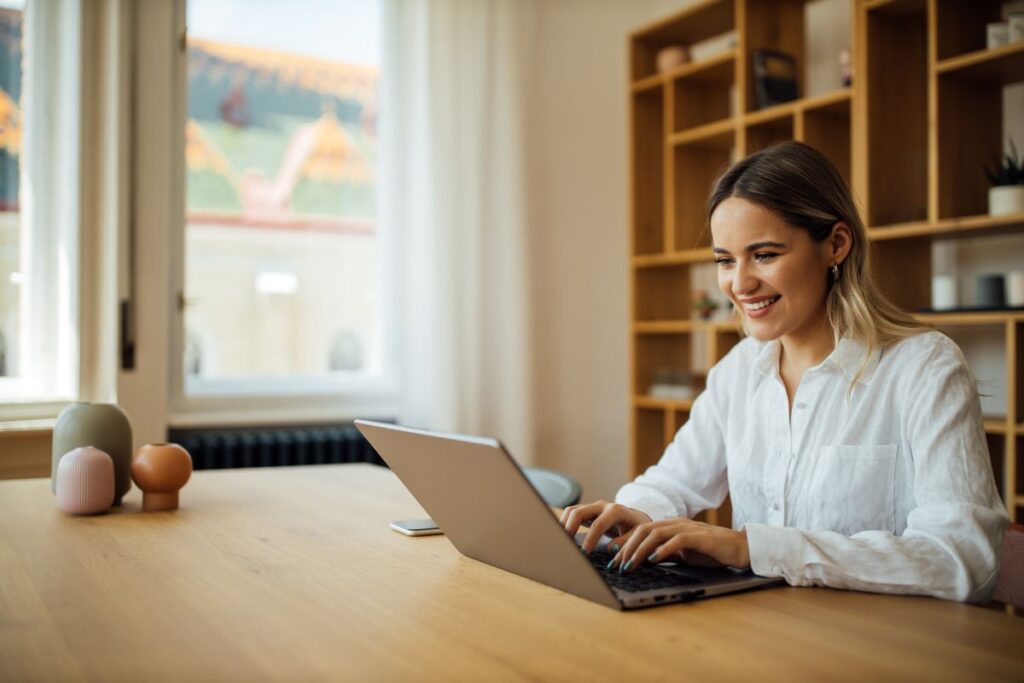 A woman works from home. She smiles at her laptop as she types.