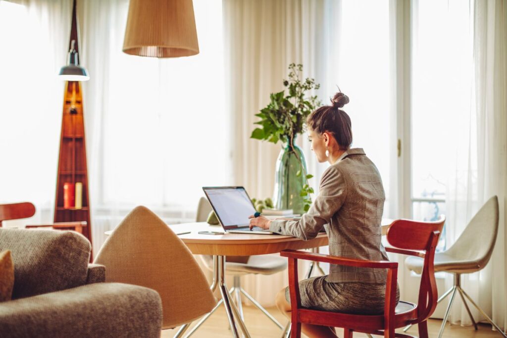 A woman works remotely. She is sat at her dining table. She types on her laptop. Her hair is in a bun.